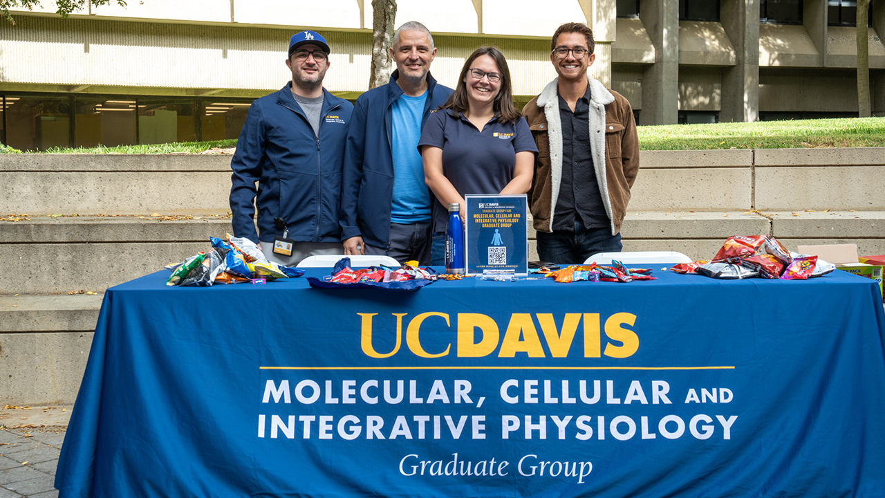 Four smiling individuals stand behind a table covered with a UC Davis tablecloth that reads 'Molecular, Cellular and Integrative Physiology Graduate Group.' The table is set up outdoors with various snacks and information materials, including a QR code sign, on display.