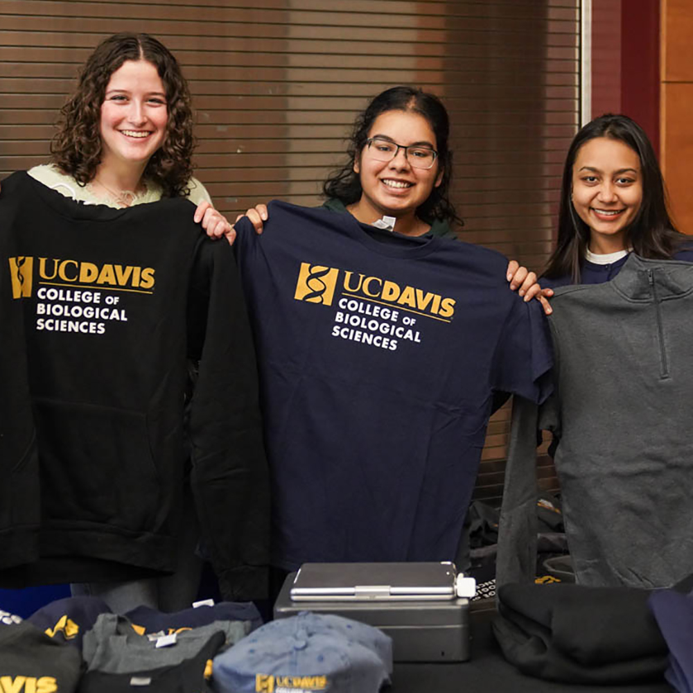 Three smiling students holding up UC Davis College of Biological Sciences apparel, including a sweatshirt and a t-shirt, at a merchandise table.