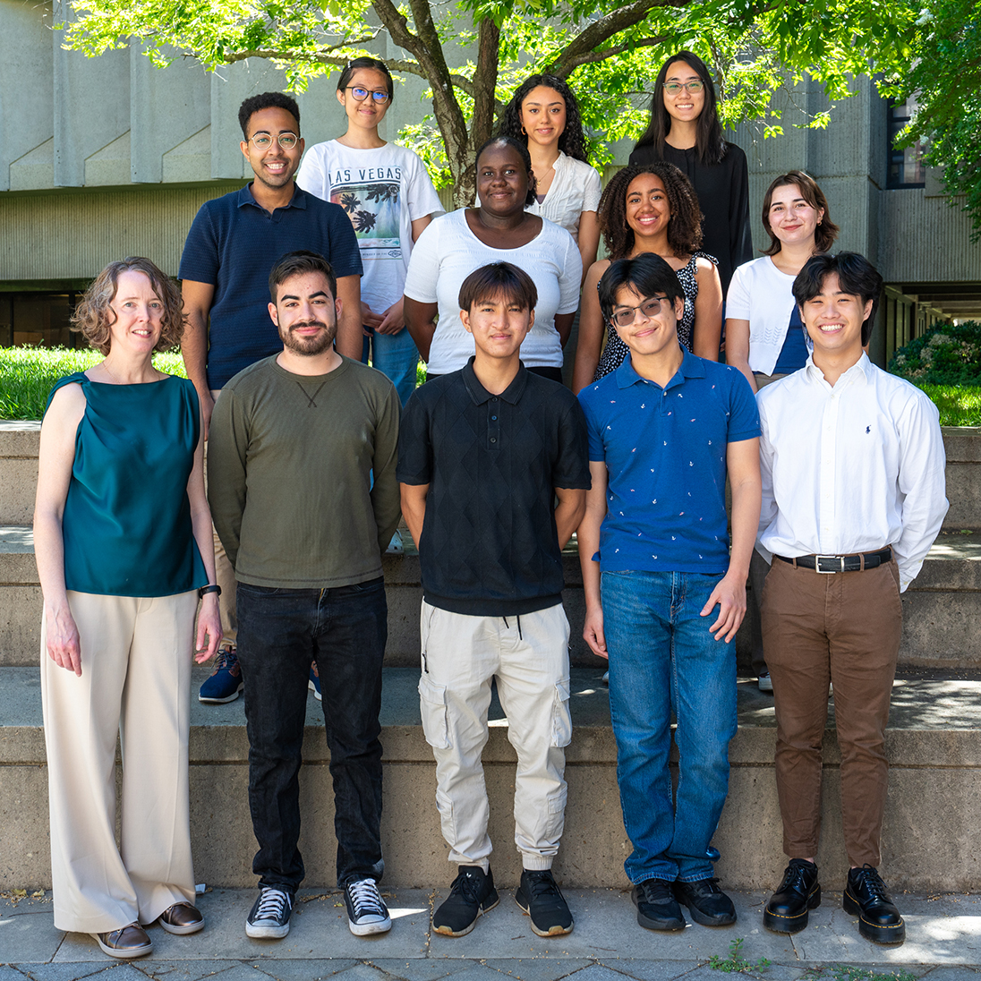 Group photo of a diverse group of students and a faculty member standing outdoors on stone steps, with a tree and campus building in the background.