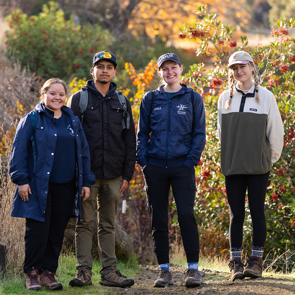 Four individuals in outdoor attire stand on a dirt path surrounded by vibrant autumn foliage, smiling at the camera during a nature outing.