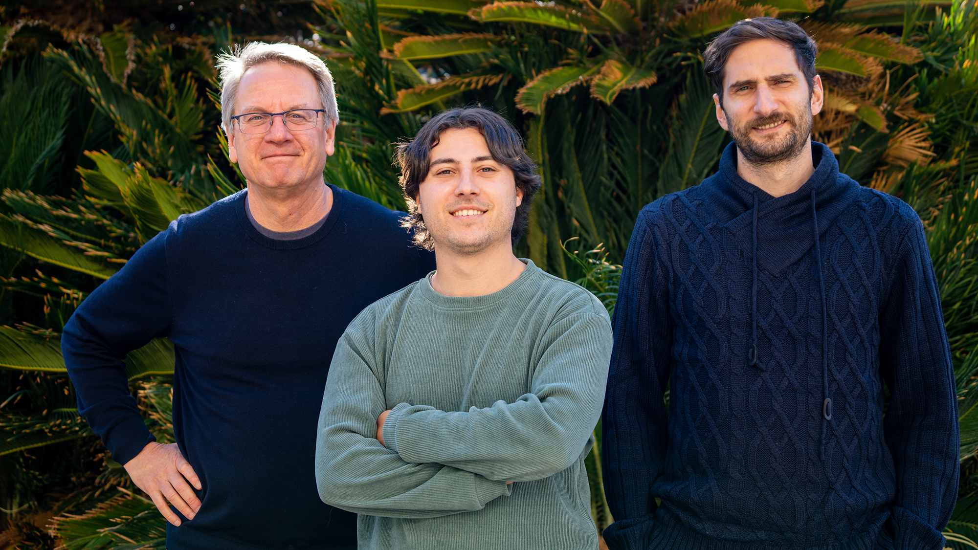 **Alt Text:** Three men stand outdoors in front of lush green foliage, smiling at the camera.