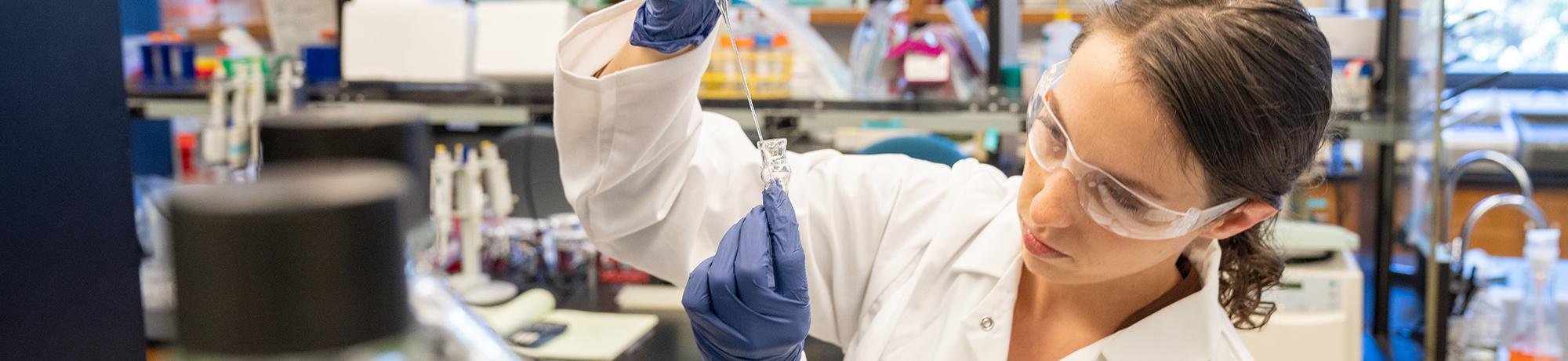 A researcher in a lab coat and safety goggles carefully pipetting a liquid into a vial, with laboratory equipment and shelves in the background.
